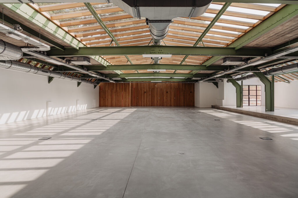 Licht industrieel interieur van een lege ruimte aan de Plantage Middenlaan met betonnen vloer, houten wand en dakconstructie met groene balken en glaspanelen.